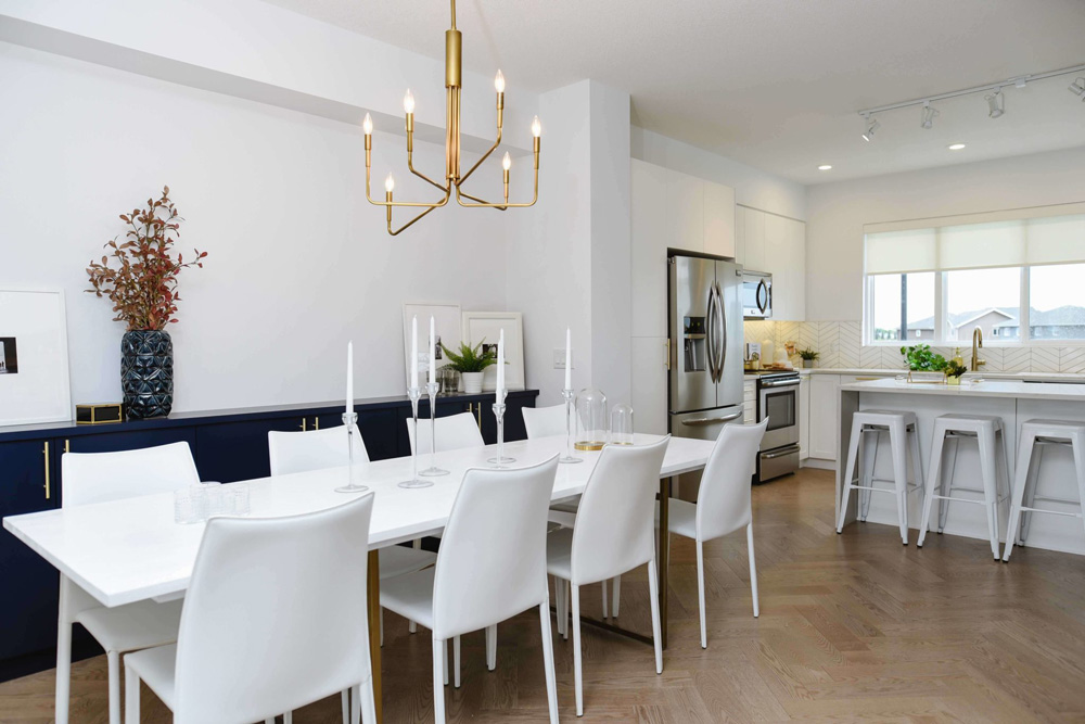 Dining room with white table and white leather chairs, connected kitchen with geometric white tile and white stools, hardwood floors and golden lights. Cobalt Beach showhome Jensen Lakes St. Albert