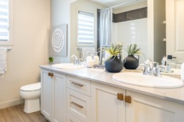 Bathroom with two sinks and white cabinets, accented with gold and hardwood floor in Pacesetter Calypso showhome in Jensen Lakes St. Albert.