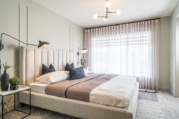 Owner's suite bedroom with large windows and white cloth bed frame, accented with matte black lighting fixtures and decor. In Jensen Lakes Pacesetter showhomes in St. Albert.